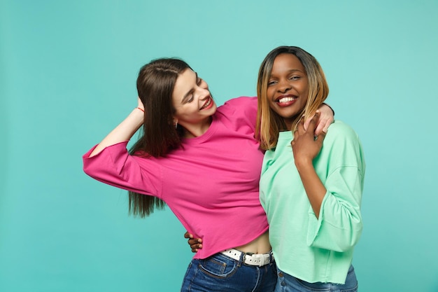 Deux jeunes femmes amis européens et afro-américains en vêtements verts roses debout posant isolés sur fond de mur bleu turquoise, portrait en studio. concept de style de vie des gens. maquette de l'espace de copie.
