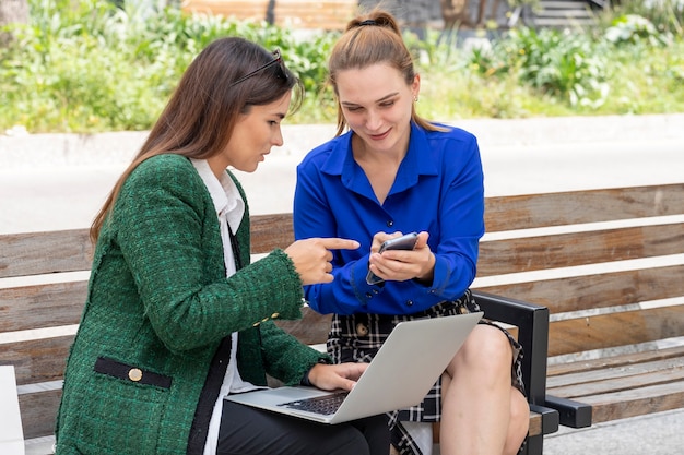 Deux jeunes femmes assises sur un banc de rue travaillant sur leur ordinateur portable pointant vers l'écran