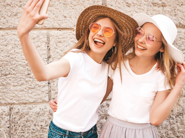 Deux jeunes femmes blondes hipster souriant dans des vêtements de t-shirt blanc d'été. Filles prenant des photos d'autoportrait selfie sur smartphone.