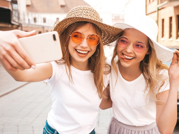 Deux jeunes femmes blondes hipster souriant dans des vêtements de t-shirt blanc d'été. Filles prenant des photos d'autoportrait selfie sur smartphone.