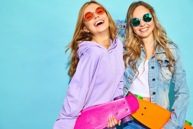 Deux jeunes femmes blondes souriantes élégantes avec des planches à roulettes penny. femmes en vêtements de sport hipster d'été posant près du mur bleu. Modèles positifs