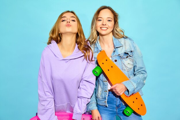 Deux jeunes femmes blondes souriantes élégantes avec des planches à roulettes penny. femmes en vêtements de sport hipster d'été posant près du mur bleu. Modèles positifs