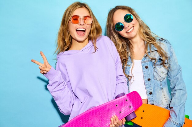 Deux Jeunes Femmes Blondes Souriantes élégantes Avec Des Planches à Roulettes Penny. Modèles En Vêtements De Sport Hipster D'été Posant Près Du Mur Bleu. Les Femmes Positives Deviennent Folles