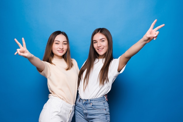 Deux jeunes femmes montrant les doigts faisant signe de la victoire isolé sur mur bleu. Numéro deux.