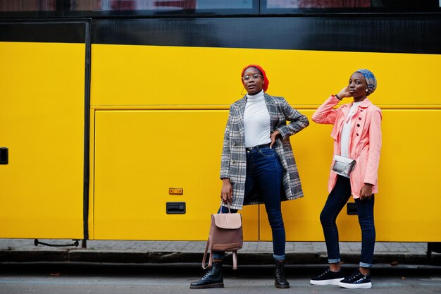 Deux jeunes femmes musulmanes africaines grandes et minces attrayantes à la mode moderne en hijab ou turban foulard et manteau posés contre le bus jaune