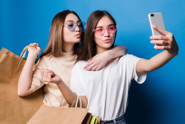 Deux jeunes femmes prennent selfie au téléphone avec des sacs en papier colorés isolés sur un mur bleu. Concept pour les ventes en magasin.