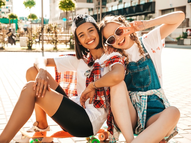 Deux jeunes filles belles souriantes avec des planches à roulettes penny colorées. Femmes en vêtements d'été hipster assis dans le fond de la rue. Modèles positifs s'amusant et devenant fous. Montrant le signe de la paix