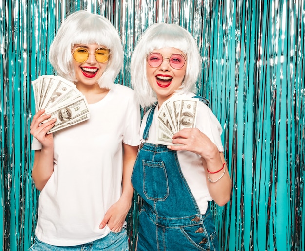 Photo gratuite deux jeunes filles sexy hipster en perruques blanches et lèvres rouges.belles femmes détenant des dollars en mains d'été dépenser de l'argent