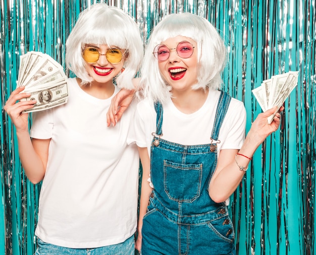 Deux jeunes filles sexy hipster en perruques blanches et lèvres rouges.Belles femmes détenant des dollars en mains d'été dépenser de l'argent