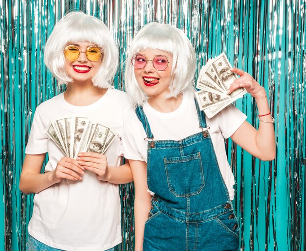 Photo gratuite deux jeunes filles sexy hipster en perruques blanches et lèvres rouges.belles femmes détenant des dollars en mains d'été dépenser de l'argent