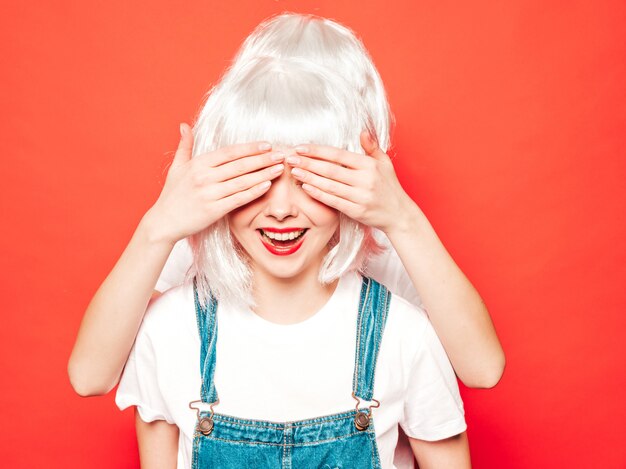 Deux jeunes filles sexy hipster en perruques blanches et lèvres rouges.Belles femmes à la mode dans des vêtements d'été.Modèles sans soucis posant près du mur rouge en studio.Couvrant ses yeux et étreignant par derrière