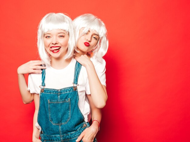Deux jeunes filles sexy hipster en perruques blanches et lèvres rouges. Belles femmes à la mode dans les vêtements d'été. Modèles sans soucis posant près du mur rouge en studio d'été en lunettes de soleil