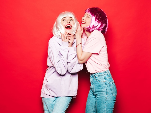 Deux jeunes filles sexy hipster en perruques et lèvres rouges. Belles femmes à la mode dans les vêtements d'été. Modèles sans soucis posant près du mur rouge en studio deviennent fous