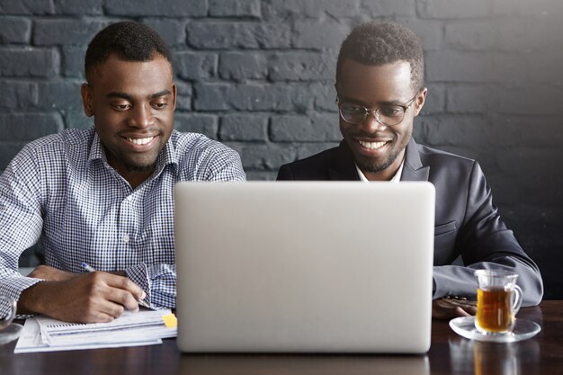 Deux jeunes hommes d'affaires afro-américains réussis joyeux assis dans l'intérieur de bureau moderne devant un ordinateur portable ouvert, regardant l'écran avec des sourires heureux, discutant des plans d'affaires et des idées