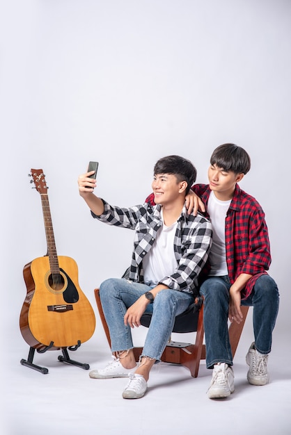 Deux jeunes hommes aimants sont assis sur une chaise et prennent un selfie depuis un smartphone.