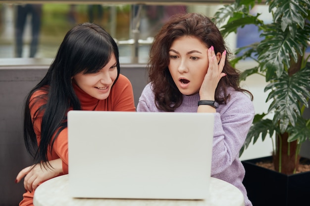 Deux jolies femmes avec un ordinateur portable blanc assis au café, regardant l'écran du cahier avec des expressions faciales étonnées, voient quelque chose d'intéressant tout en se reposant dans un café.