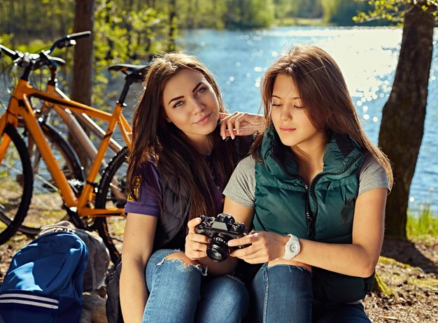 Deux jolies filles brunes utilisant un appareil photo compact et se relaxant après la balade à vélo sur la côte sauvage de la rivière.