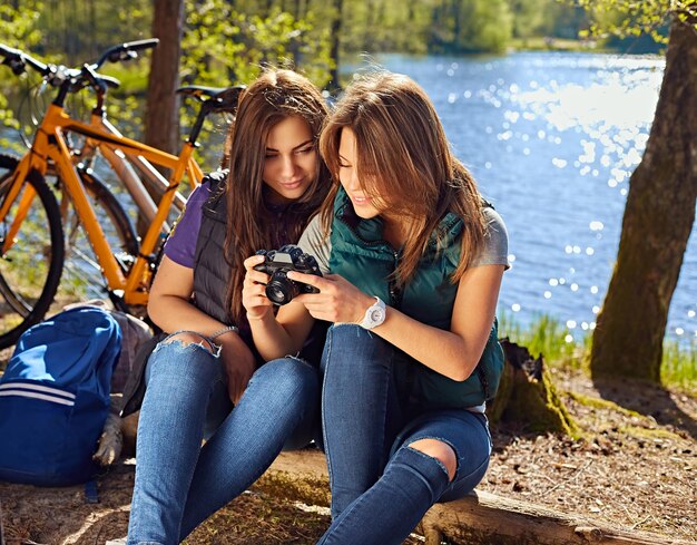 Deux jolies filles brunes utilisant un appareil photo compact et se relaxant après la balade à vélo sur la côte sauvage de la rivière.