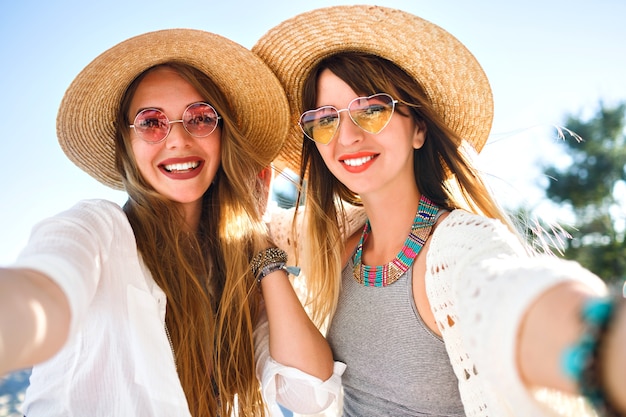 Photo gratuite deux jolies meilleures amies faisant des selfies sur la plage, des couleurs estivales claires et lumineuses, des chapeaux et des lunettes de soleil boho chic, des bijoux à la mode et un maquillage naturel, des vibrations d'amitié positives.