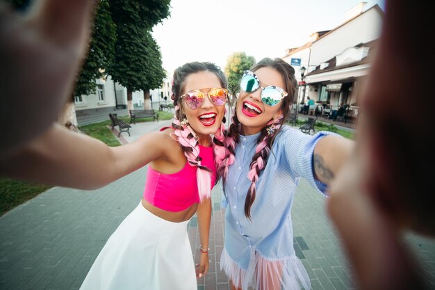 Deux meilleures copines à la mode et belles dans des verres, posant en train de faire le moi général, montrant à chacun son style de vie, passent du temps joyeusement. Les femmes en lunettes de soleil Reposez-vous dans la ville. Week-end.