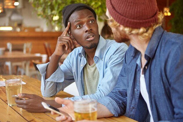 Deux meilleurs amis ou camarades d'université buvant de la bière et utilisant des gadgets électroniques au pub: un homme afro-américain parle à son ami caucasien méconnaissable, le regardant en état de choc et d'incrédulité