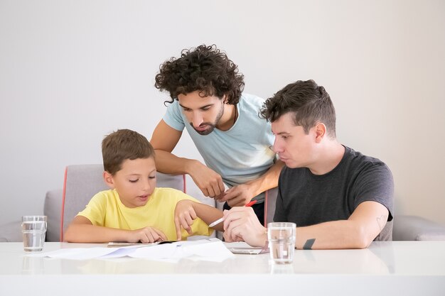 Deux papas aidant un garçon concentré avec la tâche à la maison de l'école, assis à table avec des papiers, lisant le manuel ensemble, pointant le doigt sur la page. Concept de famille et de parentalité