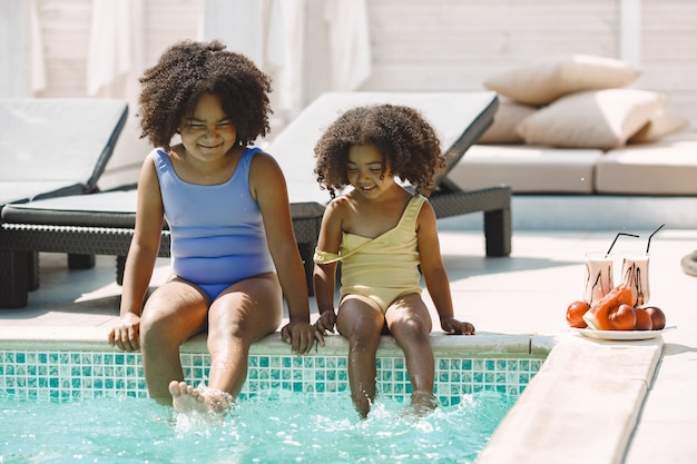 Photo gratuite deux sœurs multiraciales bouclées assises près de la piscine. filles portant des maillots de bain bleu et jaune