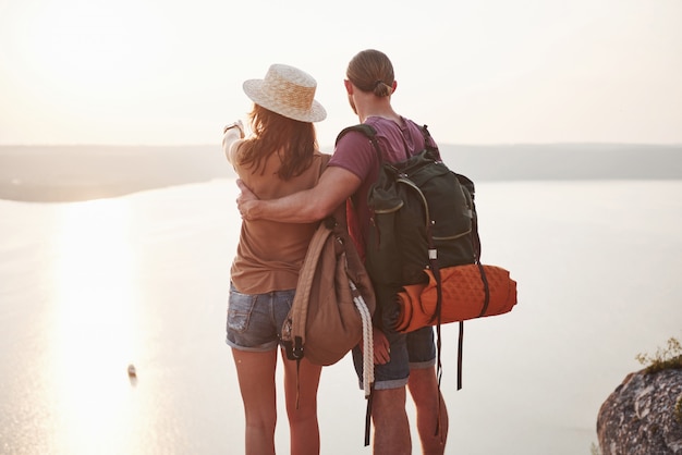 Deux touristes hommes et femmes avec des sacs à dos se tiennent au sommet de la montagne et profitent du lever du soleil.