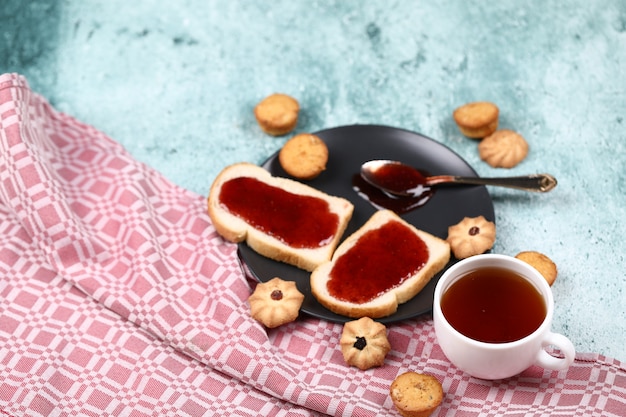 Photo gratuite deux tranches de pain grillé avec de la confiture rouge dans une assiette noire avec des biscuits autour et une tasse de thé blanc sur une table en pierre bleue.