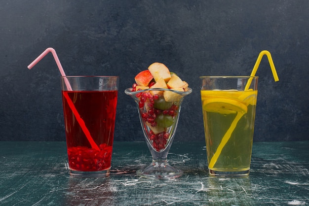 Deux verre de cocktails avec des pailles et un verre de fruits mélangés sur une table en marbre.