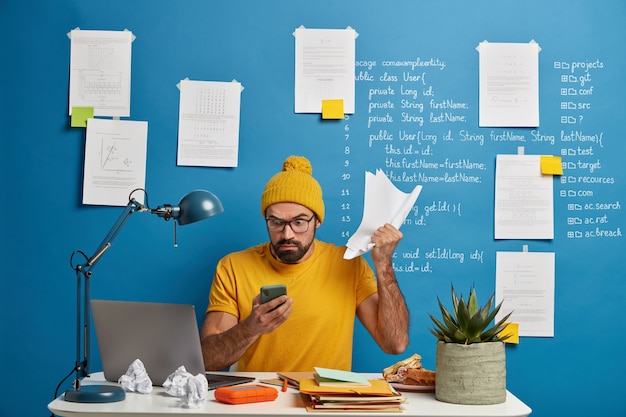 Photo gratuite le développeur informatique travaille à distance avec des documents papier, vérifie les informations via téléphone portable dans la base de données, s'assoit dans un espace de coworking