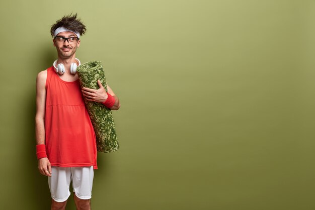 Devenir plus fort chaque jour. Un sportif réfléchi vient dans la salle de sport pour faire de l'exercice, tient un karemat enroulé, porte des vêtements de sport et des écouteurs, se tient contre un mur vert, copie un espace pour le texte.