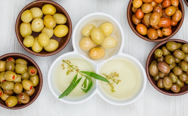 Différentes olives avec des feuilles et de l'huile d'olive dans une argile et des bols blancs sur bois blanc, gros plan.