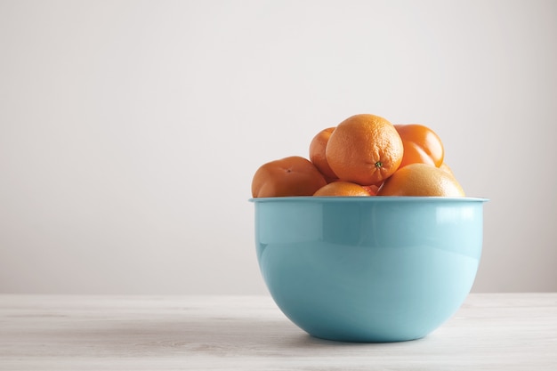 Différents fruits et agrumes dans un grand bol bleu métallique devant un mur blanc sur une table en bois blanche de côté