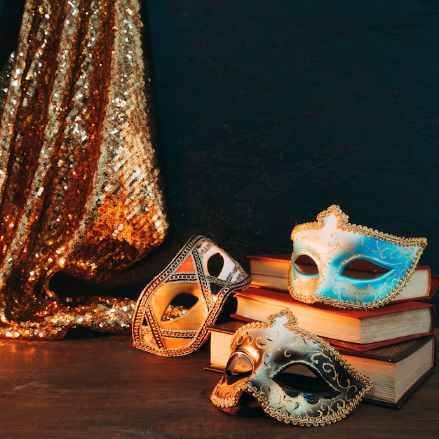 Photo gratuite différents types de masque de carnaval sur une pile de livres avec un tissu paillettes paillettes sur un bureau en bois