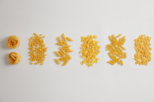 Différents types de pâtes crues crues sur fond blanc. Les nids de pâtes, farfalle, tripolini, penne, fusilli peuvent être utilisés pour les sauces ou les plats. Variété de produits italiens. Formes variées. Concept alimentaire