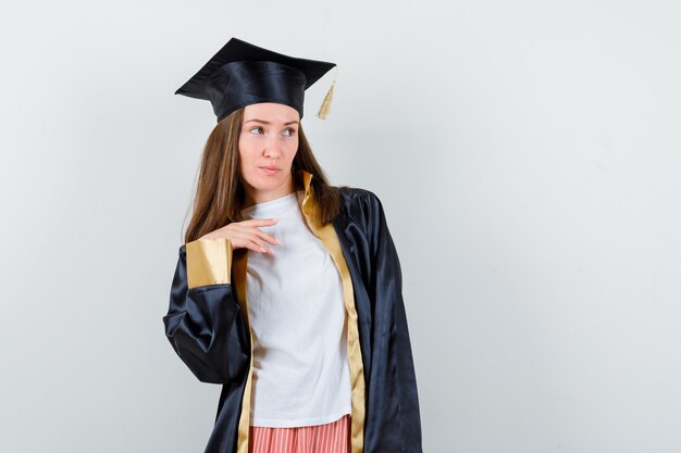 Diplômée féminine posant tout en regardant ailleurs en tenue académique et à la recherche concentrée. vue de face.