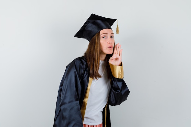 Diplômée femme disant secret derrière la main en robe académique et à la recherche de curiosité. vue de face.