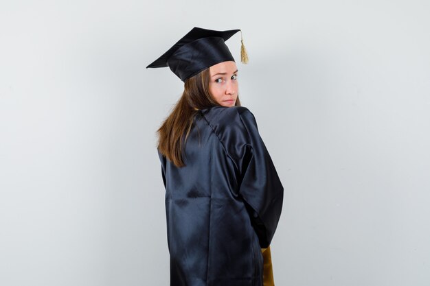 Diplômée en robe académique regardant la caméra par-dessus son épaule et à la jolie vue arrière.