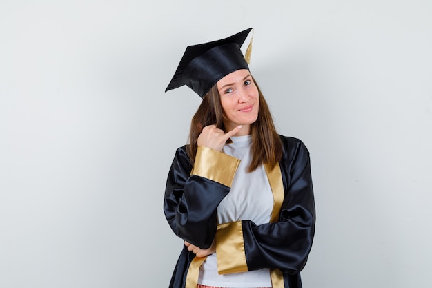 Diplômée de sexe féminin pointant vers le coin supérieur droit en uniforme, vêtements décontractés et espoir, vue de face