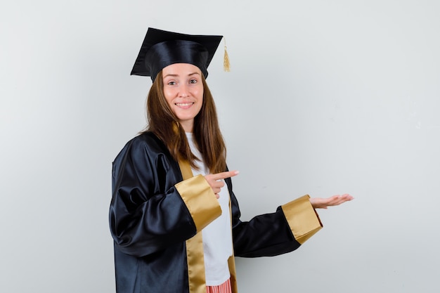 Diplômée de sexe féminin pointant vers la droite, étalant la paume de côté en tenue académique et regardant joyeuse. vue de face.