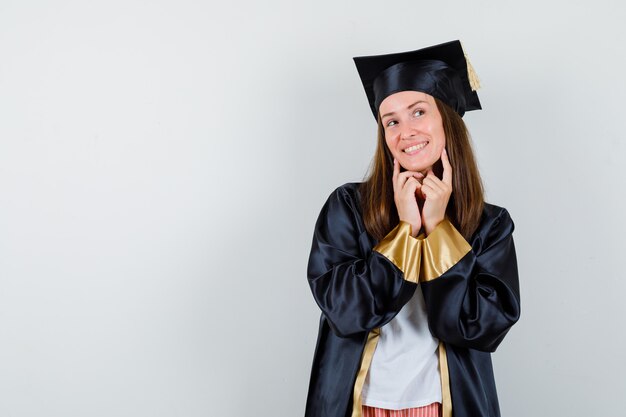 Diplômée en tenue académique en gardant les doigts sur les joues et à la rêveuse, vue de face.