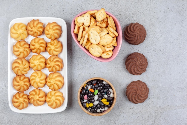 Photo gratuite disposition de biscuits sur et en dehors du plateau avec des bols de craquelins et de bonbons au milieu sur fond de marbre. photo de haute qualité