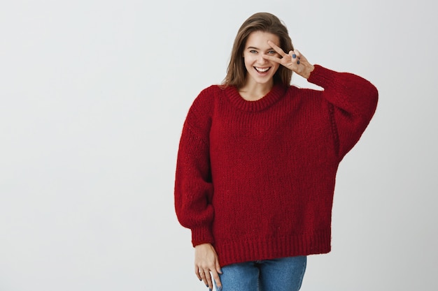 Dites "cheese. Photo de Studio de femme charmante à la mode avec un large sourire tenant une pancarte v au-dessus des yeux et exprimant des émotions positives et ludiques en se tenant debout. Soyez du bon côté