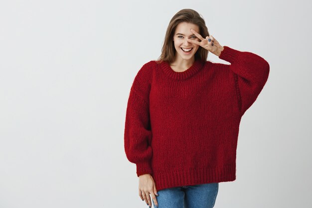 Dites "cheese. Photo de Studio de femme charmante à la mode avec un large sourire tenant une pancarte v au-dessus des yeux et exprimant des émotions positives et ludiques en se tenant debout. Soyez du bon côté