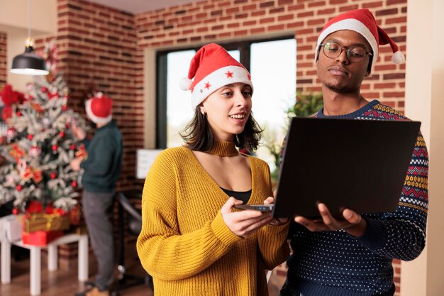 Divers collègues regardant un ordinateur dans un bureau festif, travaillant dans un espace décoré d'ornements de noël et de lumières pendant la saison des vacances d'hiver. Les gens avec un bonnet de Noel au travail la veille de Noël.