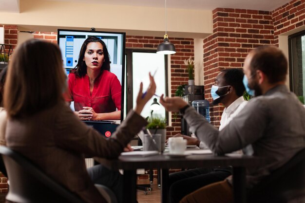 Divers gestionnaires expliquent la stratégie de l'entreprise à une femme distante lors d'une conférence de réunion par vidéoconférence en ligne travaillant dans un bureau de démarrage. Équipe d'affaires avec masque médical contre covid19