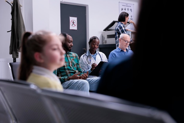 Diverses personnes attendent dans une clinique privée pendant que le médecin de l'hôpital pose des questions médicales dans la salle d'attente. Mise au point sélective sur un médecin afro-américain avec stéthoscope remplissant le formulaire pour les patients.