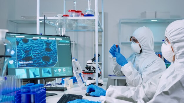 Docteur chimiste en costume ppe travaillant sur pc tandis que technicien de laboratoire utilisant un microscope. Équipe de scientifiques examinant l'évolution du vaccin avec la haute technologie pour la recherche d'un traitement contre le virus covid19