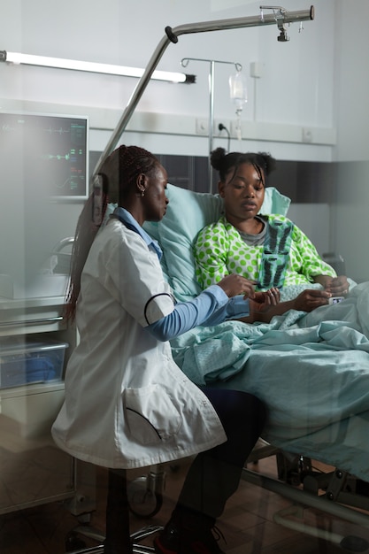 Docteur d'ethnie afro-américaine tenant une radiographie dans une salle d'hôpital. Femme afro regardant les rayons x avec un jeune patient pour la récupération du traitement. Fille noire assise dans son lit à parler au médecin